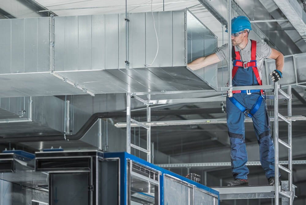 Sheet metal worker in a fall-protection harness installing rectangular ductwork from a scaffold
